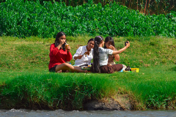 Four girls gather on riverside grass grilling skewers and chicken, with one drinking coffee and...