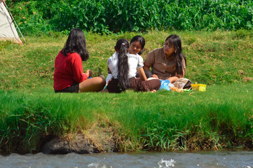 Seated together on riverside grass, four girls chat warmly as skewers of food cook over a portable...