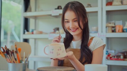 Asian woman painting ceramic art and pottery designs in her studio – happy artisan potter sitting with handmade tools, enjoying sculpting process and creative small business lifestyle