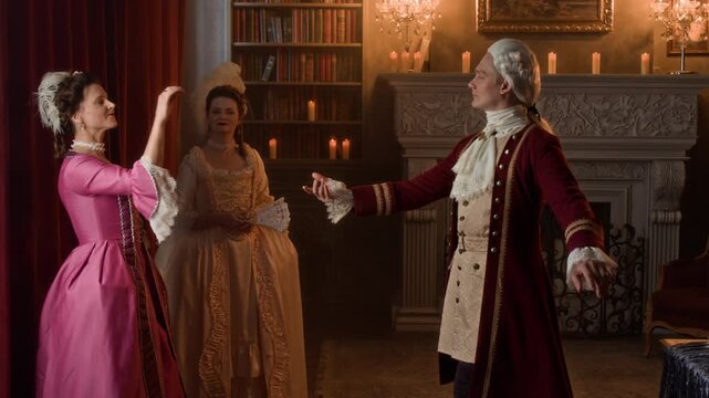 Medium full shot of polite Victorian period gentleman, wearing red frock coat, breeches, wig approaching to ladies in luxurious dresses, bowing and asking one to dance, while attending ball in palace