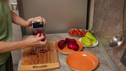 Close-up of a woman grating fresh beets on a box grater over a cutting board, vibrant red shreds falling into place. Preparation of a vegetable dish. Beets for making borscht. Close-up.