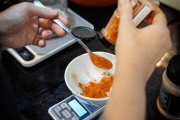 Chef's hand pours orange spices into cup on kitchen scale placed on kitchen counter.