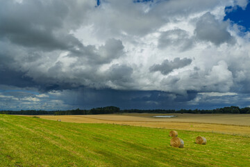 A storm is approaching over the fields, where hay bales are arranged on the fresh green grass. Dark clouds create a dramatic contrast with the peaceful countryside.