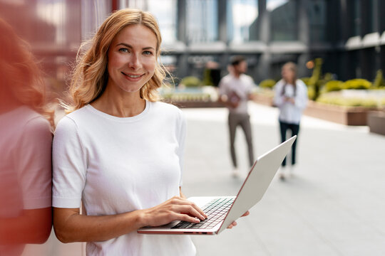 Businesswoman using laptop standing near office building, colleagues talking in background