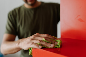 Cropped shot of housekeeper male cleaning red shelf with rag and detergent, ensuring spotless and dust-free surface in cleaning routine, close-up. Concept of domestic work, housekeeping and household.