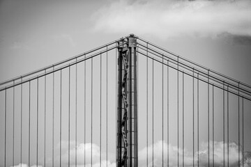 Minimalist black and white photo of a suspension bridge tower with vertical cables and clouds in the background. Architectural detail highlighting strength, symmetry, and engineering design