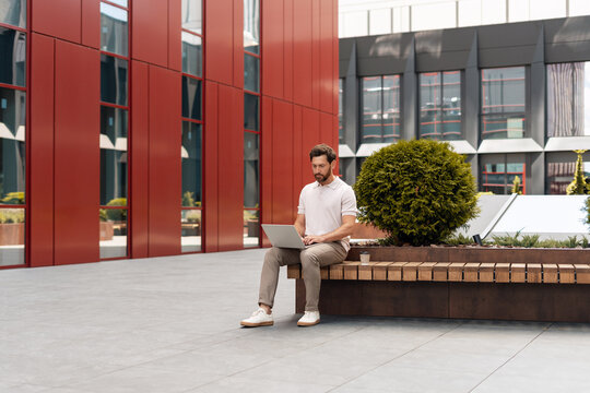 Businessman working on laptop sitting on bench in modern office district - Powered by Adobe