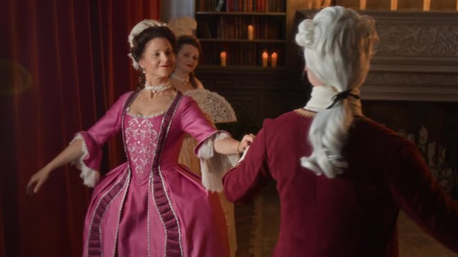Medium shot of young noble Caucasian lady in opulent pink dress and gentleman in red surcoat, jabot, wig dancing menuet together at ball in royal palace, woman with fan is watching from afar