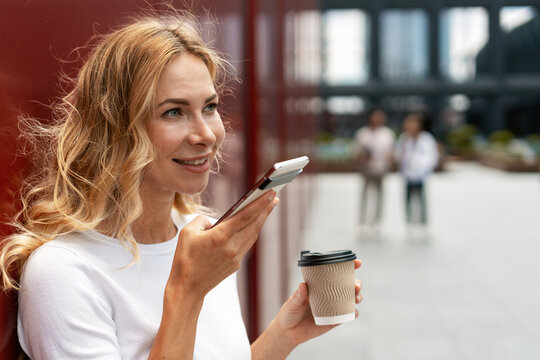 Businesswoman using smartphone voice assistant and drinking coffee outdoors