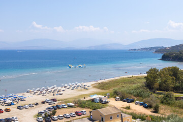 An elevated panoramic shot of a crowded Greek beach and resort area, a busy parking lot, umbrellas,...