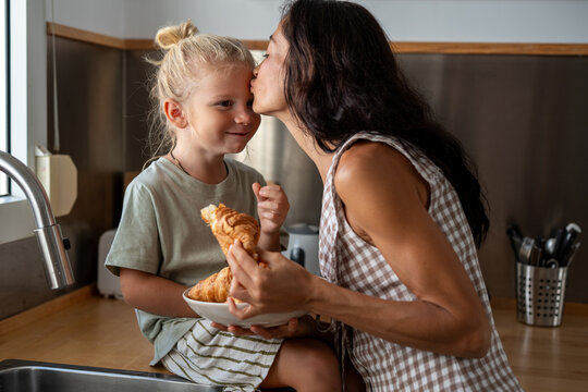 Loving mother kissing child on forehead in kitchen while holding plate with fresh croissants, sharing warmth, affection and family moment together