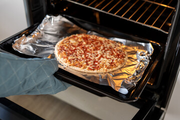Close up of homemade pizza baking in oven on tray with aluminum foil, melted cheese and tomato sauce visible