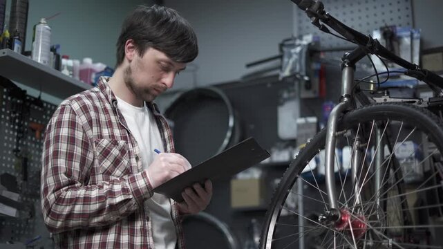 Dedicated bicycle mechanic meticulously works in workshop. He is focused and skilled, providing quality maintenance and repair services for clients bike. Male cycle fitter operating with paperwork 