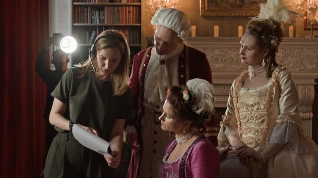Medium shot of Caucasian female movie producer and actors in 19th century costumes checking dialogues in scene script, technician setting up lights, while shooting historical drama series for TV