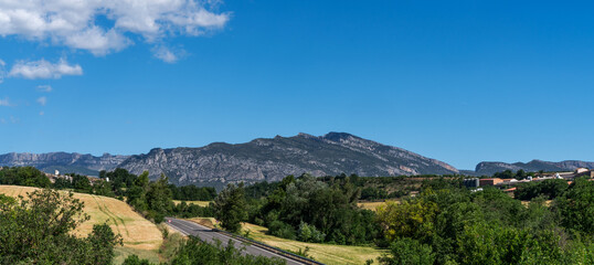 Obraz premium Rugged beauty of the Montsec mountain range in Catalonia, Spain. Scene includes rolling hills, a winding road, and a clear blue sky, creating a picturesque and tranquil landscape typical of the region