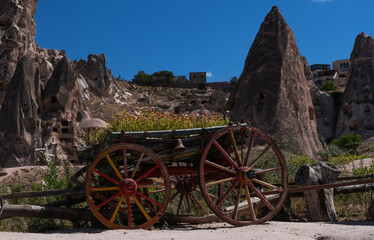 Obraz premium An old wooden cart with vibrant red and yellow wheels stands against a backdrop of unique fairy chimneys and a hill adorned with flowers under the azure sky of Cappadocia.