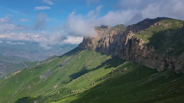 Rocky cliff wall towers above the highland of Aktoprak Pass in Kabardino-Balkaria. Sunlit grass and long shadow reveal relief and broken scree. Grand panorama ideal for travel content