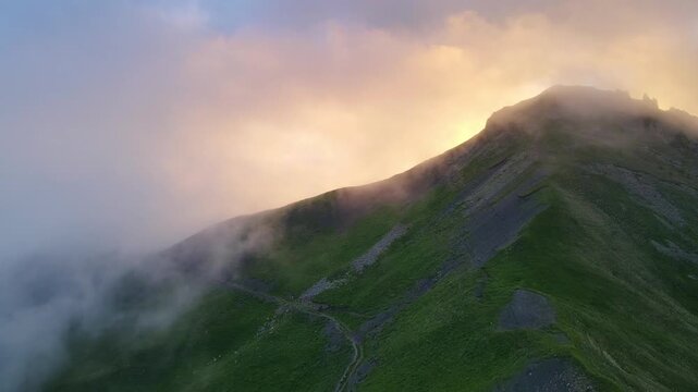 Aerial view of a foggy ridge path at Aktoprak Pass in Kabardino-Balkaria. Warm sunrise glows through mist along the crest while slopes fade below. Moody mountain atmosphere with cinematic depth