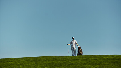 Golfer walking across green hill carrying golf bag under bright sky. Concept of endurance, sports lifestyle, recreation, training, discipline and healthy activity.