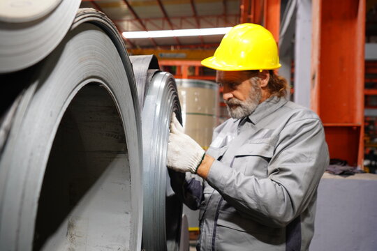Senior male technician wearing safety helmet walks with walkie-talkie to check the quality of rolled steel sheet inventory in a factory.