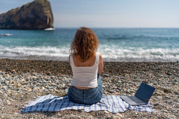 Beach Laptop Woman: Back view of woman using laptop on pebble beach, enjoying coastal views in daytime.