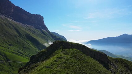 Morning light paints the green ridge of Aktoprak Pass, Kabardino-Balkaria, Russia, as low cloud drifts across deep valleys below cliffs; the drone glide reveals epic scenery and a strong travel mood