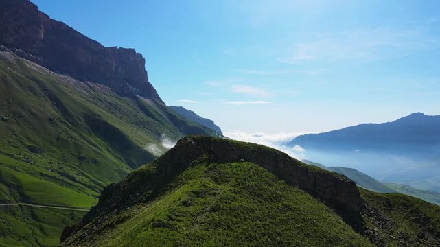 Morning light paints the green ridge of Aktoprak Pass, Kabardino-Balkaria, Russia, as low cloud drifts across deep valleys below cliffs; the drone glide reveals epic scenery and a strong travel mood