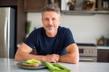 Smiling middle-aged man sitting at kitchen counter with healthy avocado toast. Mature male enjoying fresh breakfast in bright modern home interior.