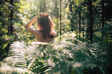 Back view of a woman standing in a lush green forest, touching her hair while sunlight filters through the trees and ferns.