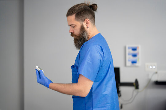 Doctor wearing blue scrubs and gloves checking pulse oximeter reading in hospital room