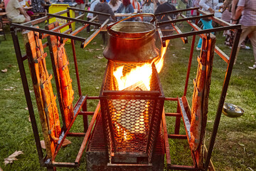 Large copper pot of fish soup and several salmon halves on grill are cooked over open fire at...