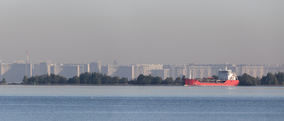 Fototapeta premium Tranquil waterfront view with red cargo ship sailing on calm water, modern high-rise buildings in haze, and green shoreline vegetation.
