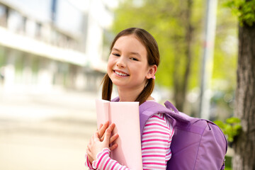 Smiling young schoolgirl outdoors holding books and wearing a backpack, enjoying a bright day in...