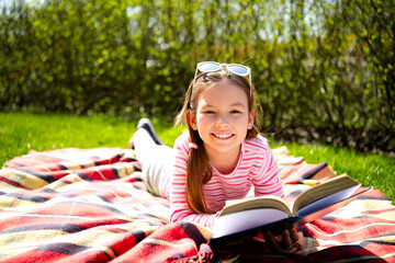 Cheerful young girl reading a book on a colorful blanket outdoors on a sunny day, enjoying the warm weather