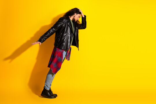 Attractive stylish man with long dark hair poses confidently in a leather jacket against a yellow background exuding fashion
