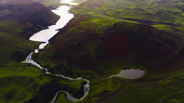 Aerial view of green volcanic terrain with a crater, winding river, reflective lakes at sunset. Smooth camera panning motion in Iceland.