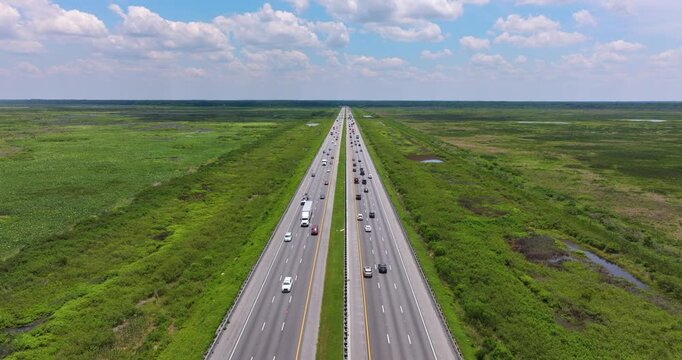 Bright cloudless sky over Florida highway lined with swamp vegetation and straight road lanes filled with cars and trucks.