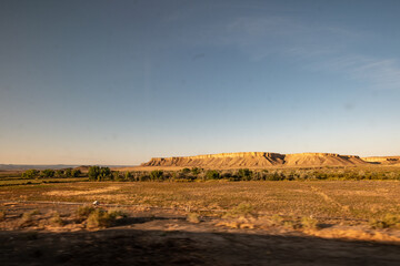 Fototapeta premium Golden light bathes a vast arid plain dotted with scrub, a distant sandstone mesa and green riparian ribbon beneath an expansive blue sky.