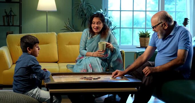 Indian kid playing carrom with grandparents in modern home indoors, elderly grandmother and grandfather enjoying board game with child, family bonding, traditional Indian lifestyle, fun, joy
