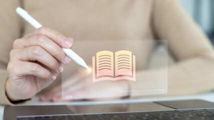 Hand using a stylus pen on a laptop with a digital open book icon hologram, concept of e-learning, online education, digital library, virtual classroom, and smart learning technology.
