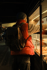 Side view of a man in a red vest and beanie at a bakery counter, looking at pastries inside the glass case while carrying a large backpack in the evening light