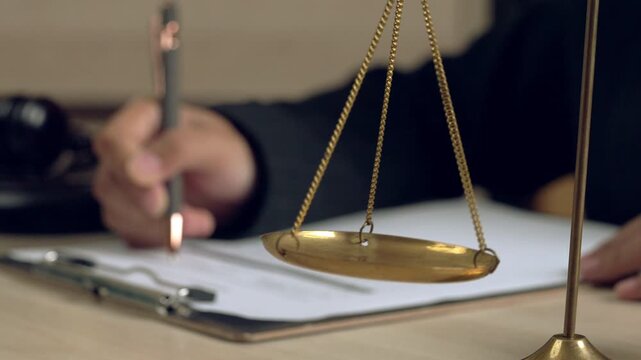 Focus on golden justice scale with blurred hand signing legal contract in background, symbolizing law, justice, legal decision, and agreement.