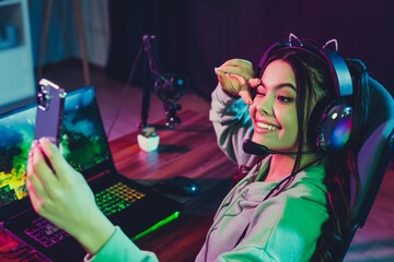 Young woman gaming and live streaming from home using laptop and mobile phone in vibrant neon light...