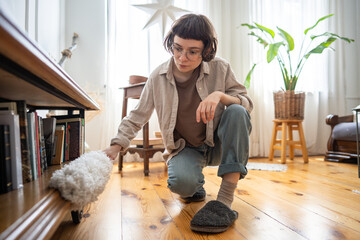 Young housewife crouching slightly, wiping shelves and low surfaces with precision cleaning tool in tidy interior. Dust removal, floor level furniture cleaning, thorough household maintenance cleaning © DimaBerlin