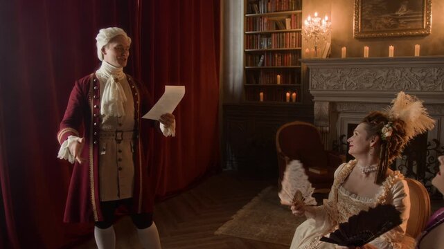 Medium full shot of young Victorian period Caucasian male aristocrat, wearing red surcoat, breeches, lace cravat, wig entertaining two royal ladies while reading aloud own romantic poems