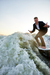 Young man surfer in classic suit rides on the wave. Happy clerk escaped from stuffy office to take up active sport. Best summer leisure after routine work. Film grain effect. Sun flare.