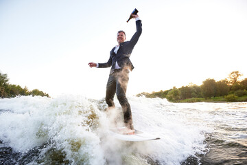 Young man in classic suit with champagne bottle rides a wakeboard on the lake near city. Happy clerk escaped from a stuffy office to take up active sport. Best summer leisure after routine work.