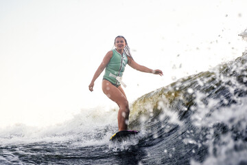 Young beautiful sporty girl in a green life jacket surfing on the wave. Happy sportswoman showily glides on the board. Active lifestyle, healthy hobby. Film grain effect.