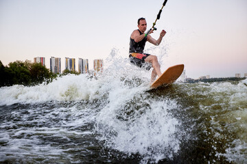 Young athletic man vigorously rides a wakeboard holding halyard. Sportsman in black swimsuit glides spectacularly along the wave like a real surfer. Active lifestyle and extreme sports.