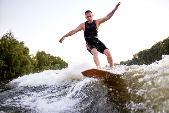 Young athletic man vigorously rides a wakeboard. Handsome guy in black swimsuit glides spectacularly along the wave like a real surfer. Active lifestyle and extreme sports. Summer watersports hobby. - Powered by Adobe
